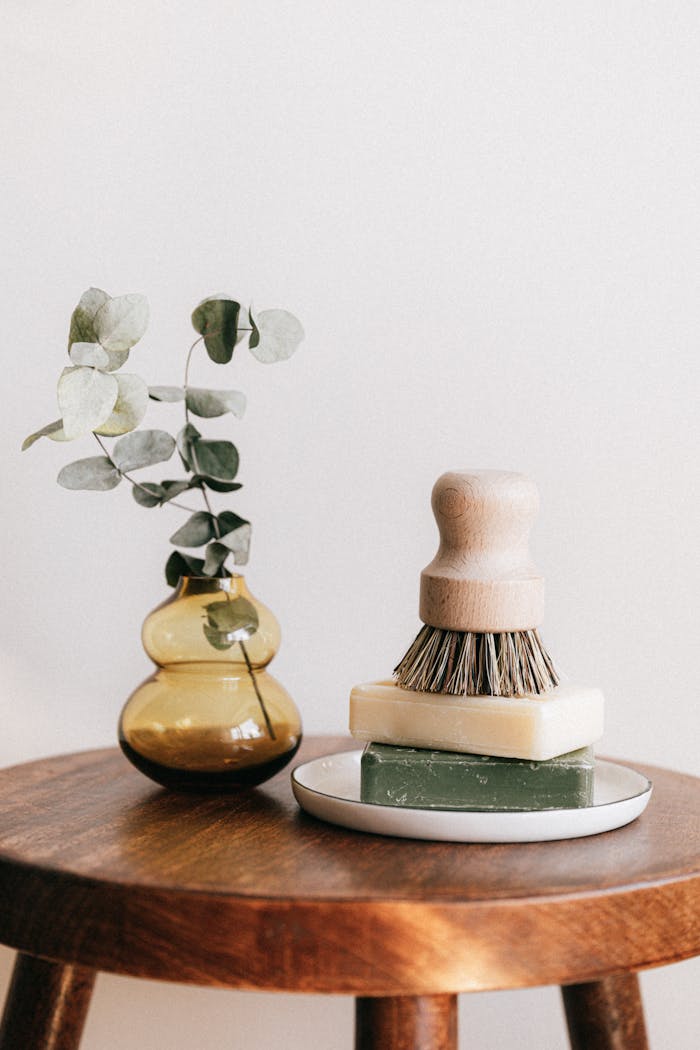 portfolio-03 Composition of shaving brush placed on organic soaps on ceramic white holder near yellow transparent vase with plant on wooden round table against white wall